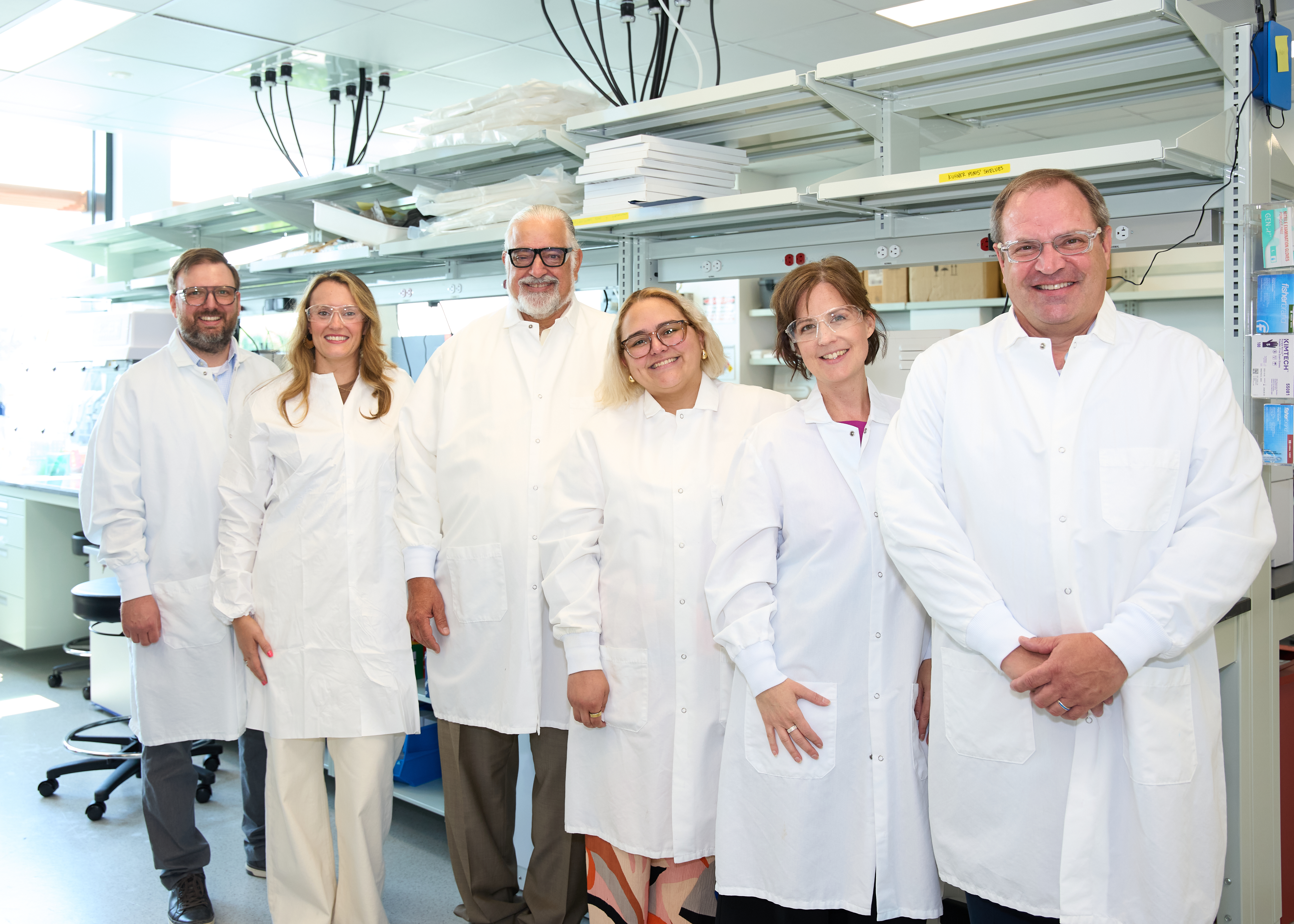 Six adults stand smiling while wearing white lab coats in front of medical research laboratory equipment.