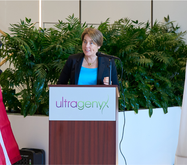 A woman with short brown hair stands behind a podium with an Ultragenyx logo, talking into a microphone.